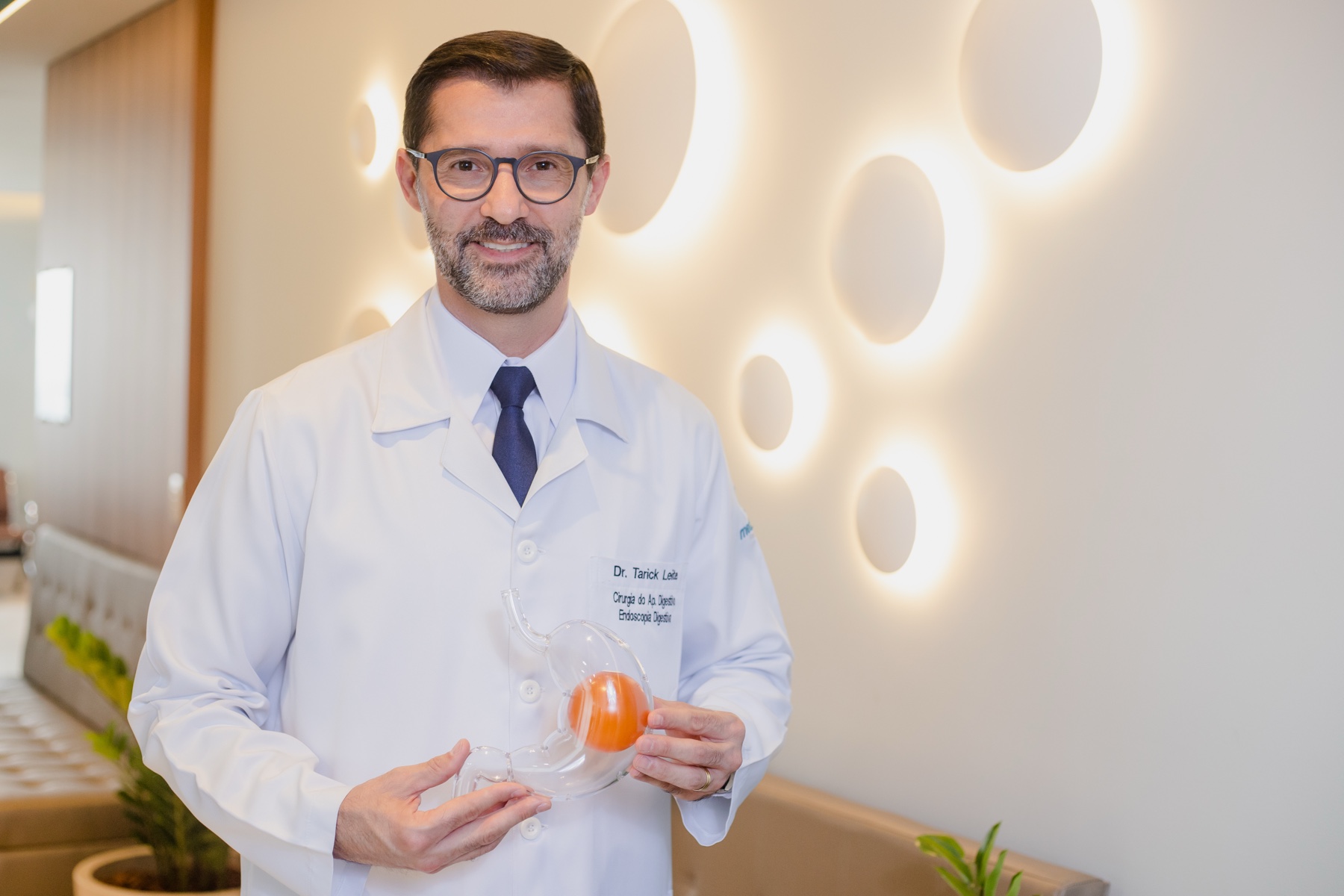 Dr. Tarick Leite de jaleco apresentando um modelo do estômago